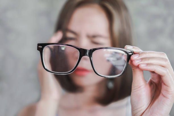 Woman holding glasses with blurry lenses, squinting, appears to have a headache.