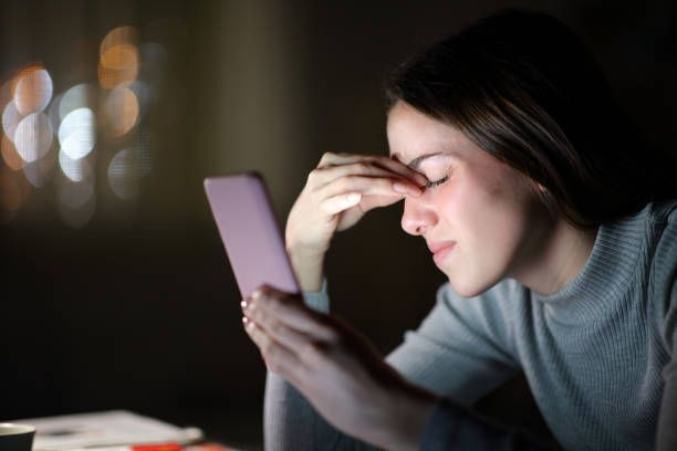 Woman in dim light holds a phone, rubbing her tired eyes.