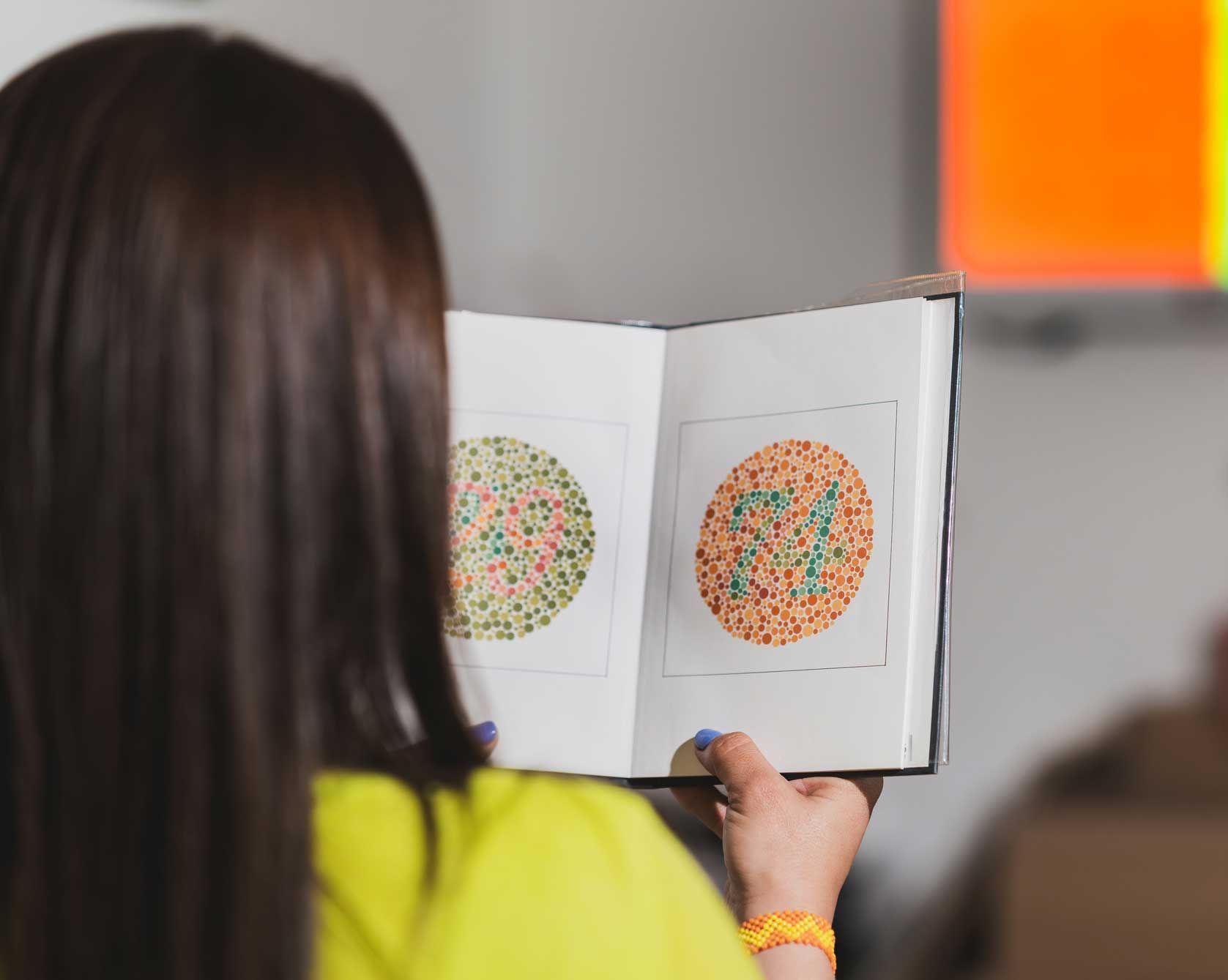 Woman holding a color blindness test book, displaying colored dot patterns. Indoor setting.