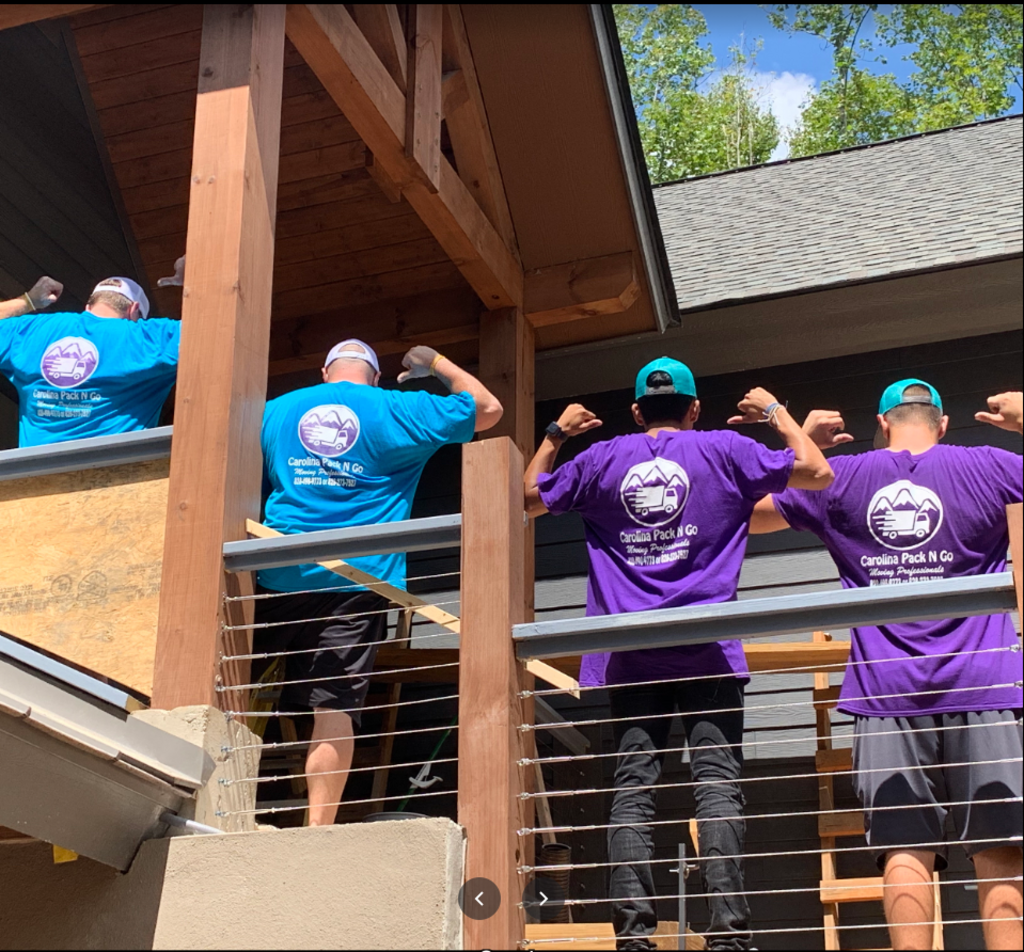 Four people flexing their biceps; two in blue shirts, two in purple shirts; standing near a wooden structure.