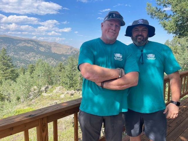 Two men in teal shirts pose on a wooden deck overlooking a mountain landscape under a blue sky.