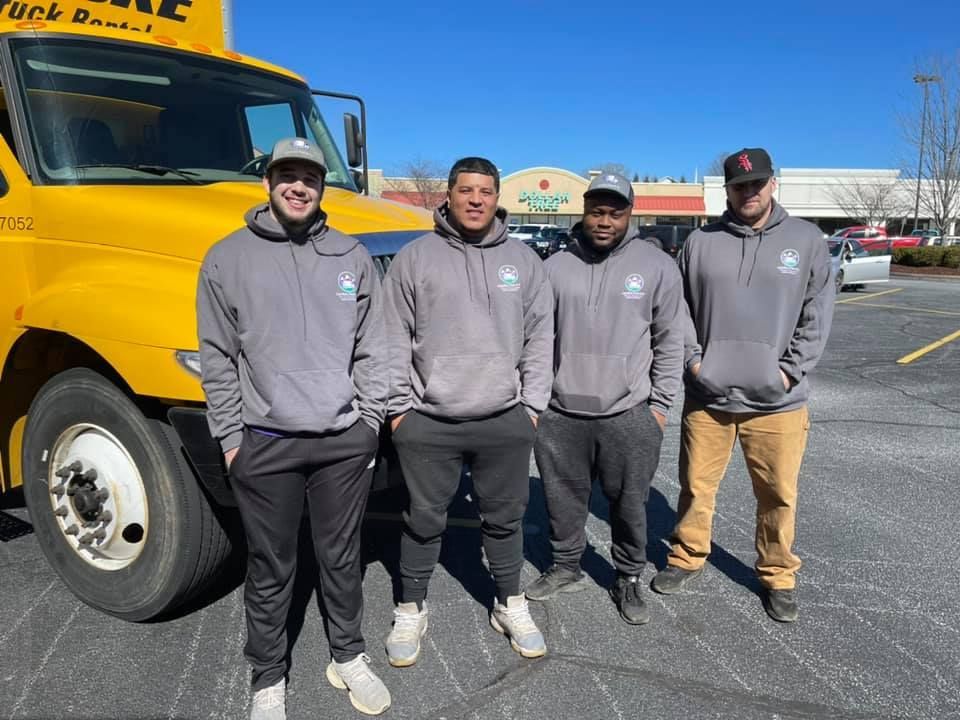 Four movers standing next to a yellow moving truck in a parking lot on a sunny day.