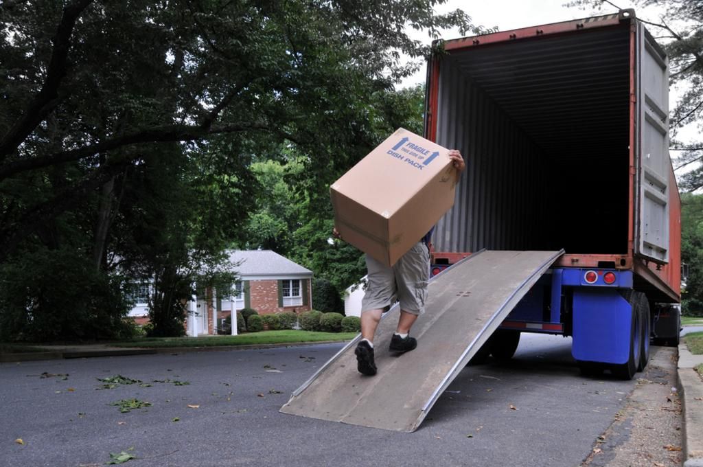 Person carrying a box up a ramp into a shipping container on a street in front of a house.