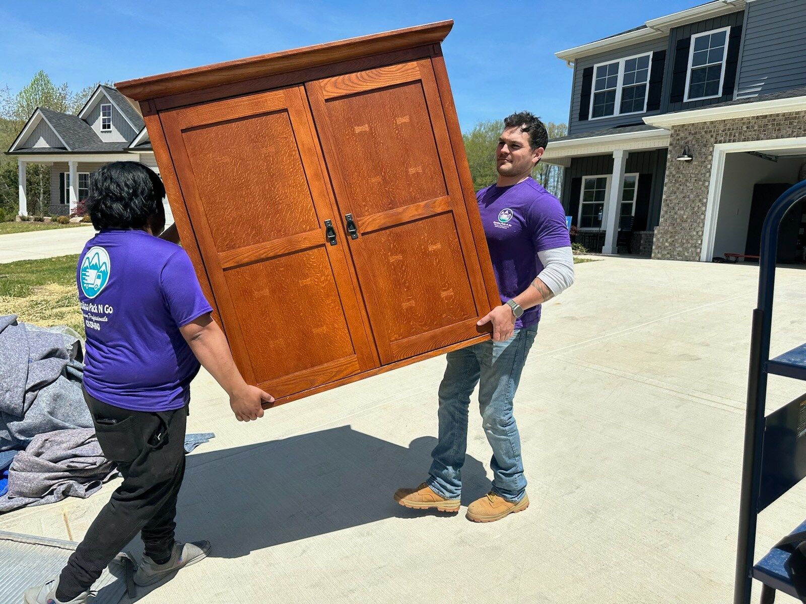 Two movers carrying a large brown cabinet from a driveway to a house.