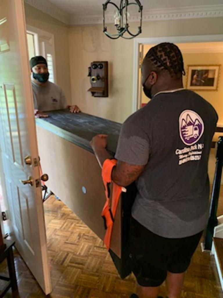 Two movers, wearing masks, carrying a large item out of a doorway; interior home with stairs and a light fixture.