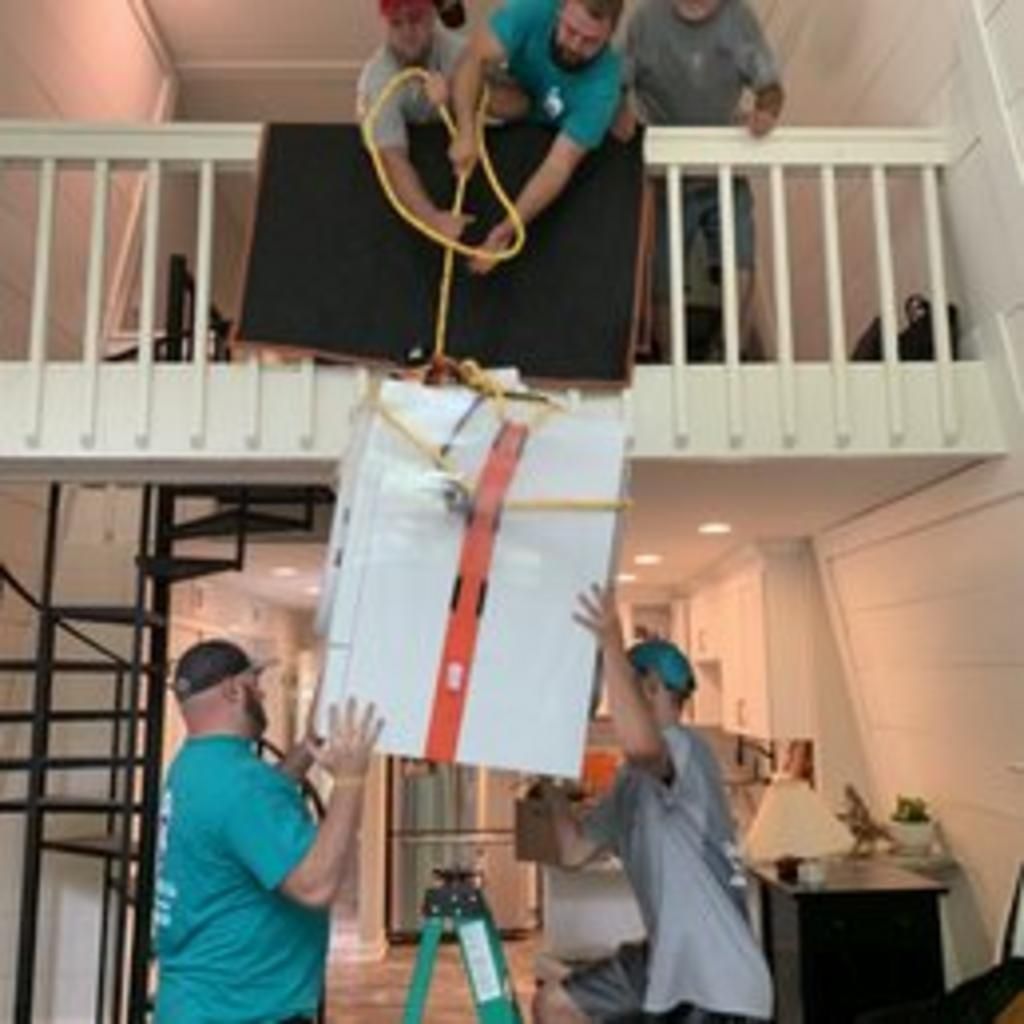 Movers lowering a refrigerator through a second-story opening with ropes. Men in teal shirts.