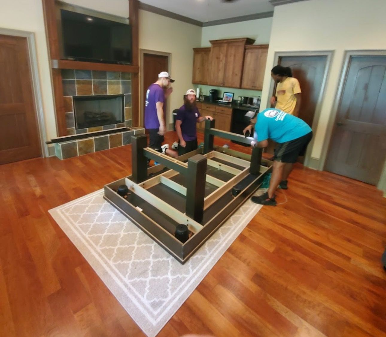 Four people assemble a dark table on a light rug in a room with wood floors.