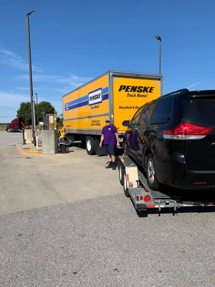 A man by a Penske truck and a car on a trailer at a gas station.