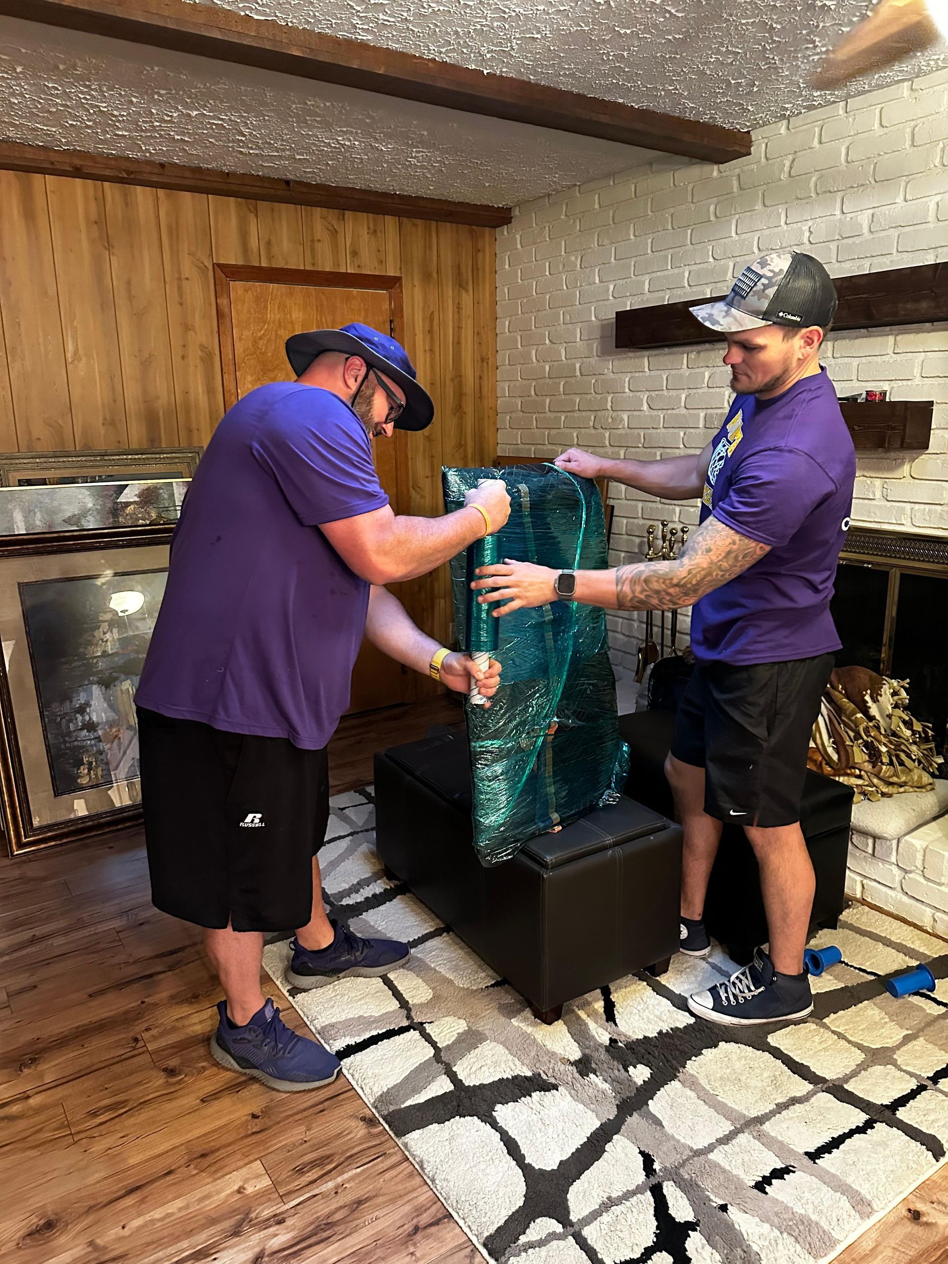 Two men in purple shirts lift a large, wrapped, blue-green object in a living room.