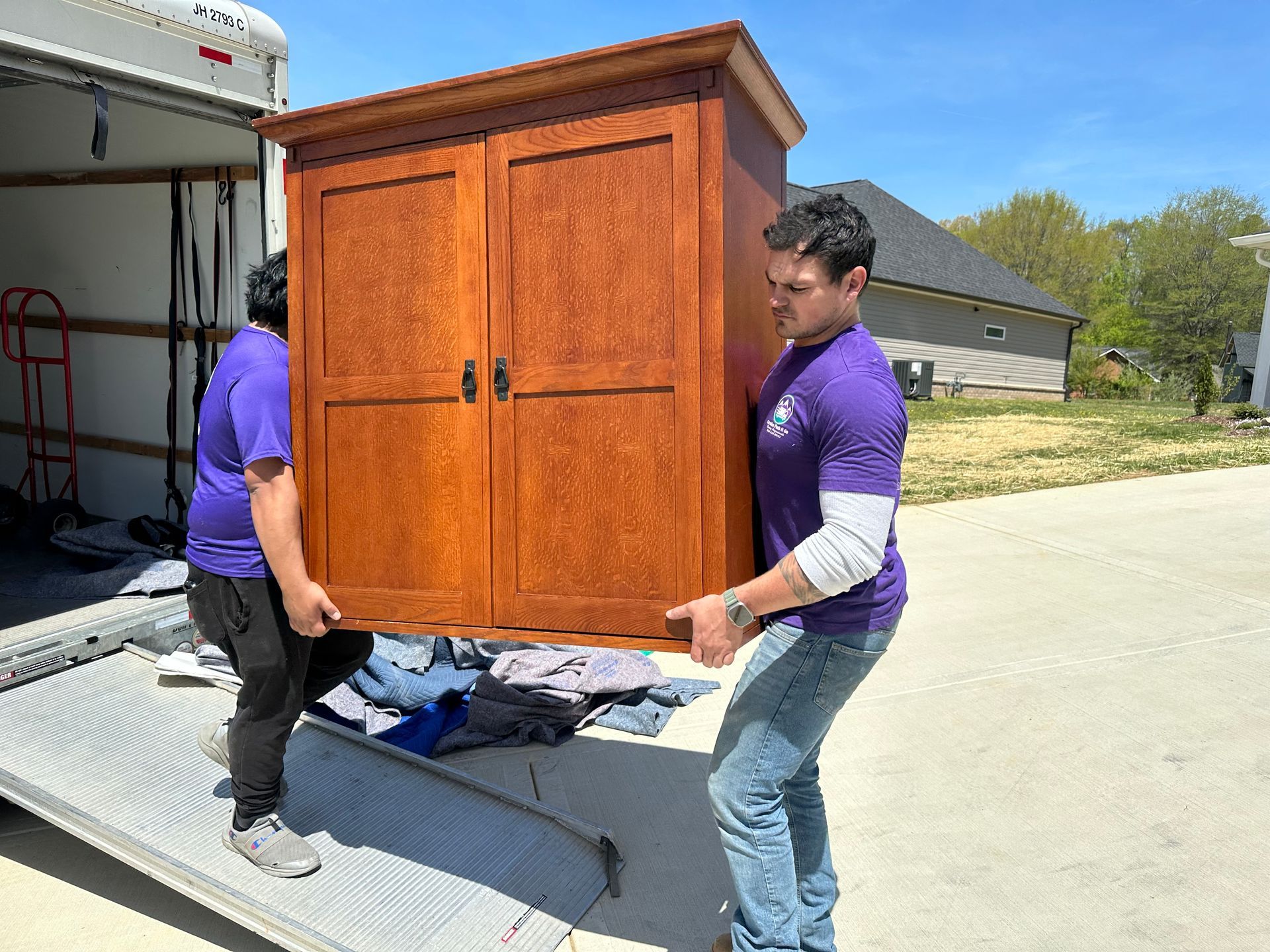 Two men in purple shirts carrying a wooden cabinet out of a truck onto a driveway.