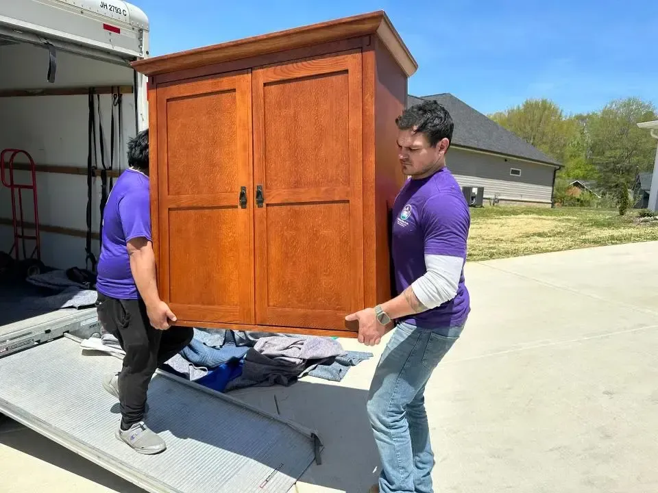 Two men in purple shirts carrying a large wooden cabinet off a moving truck. Sunny day.
