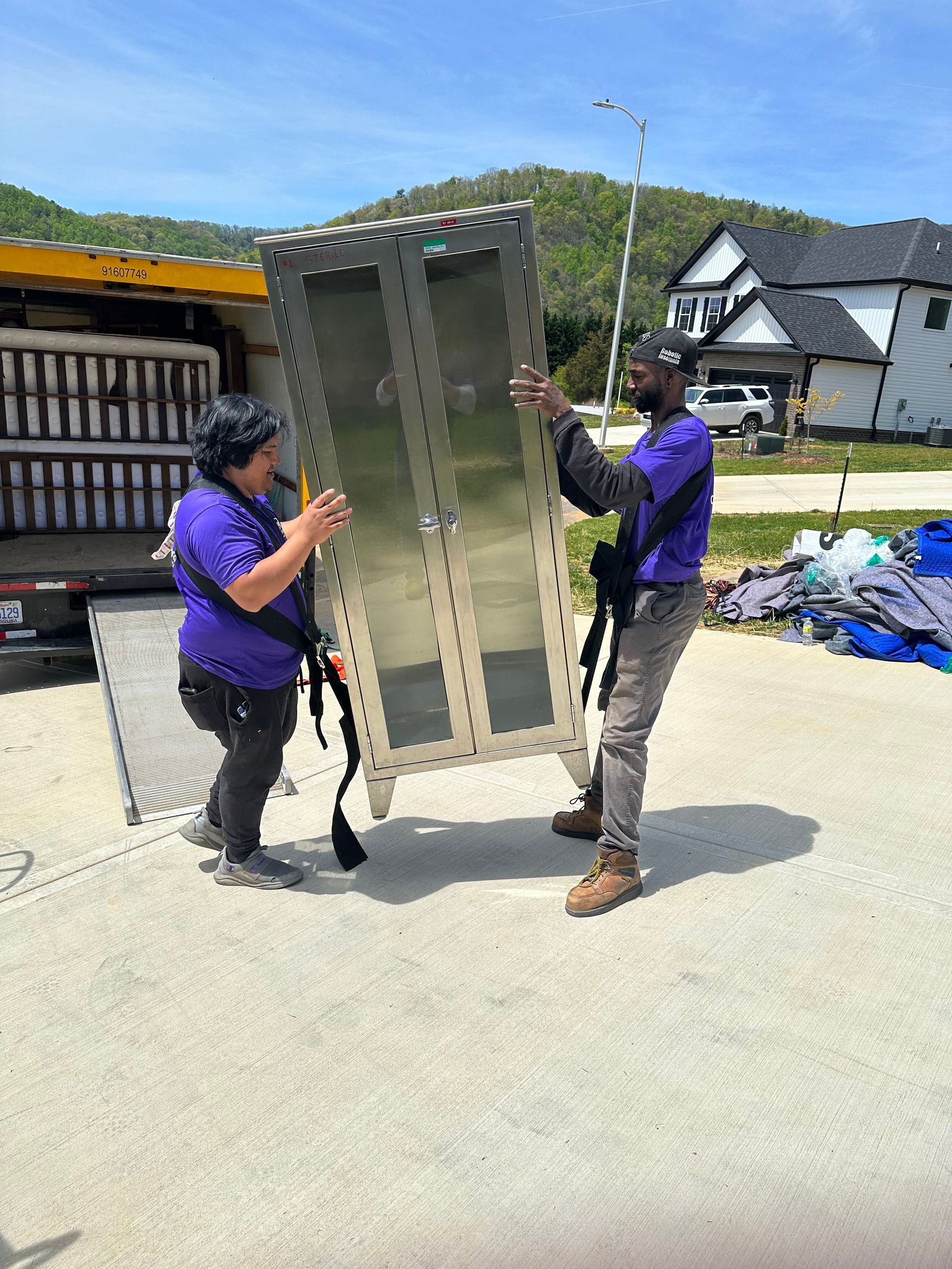 Two people in purple shirts lifting a large cabinet onto a truck outdoors.