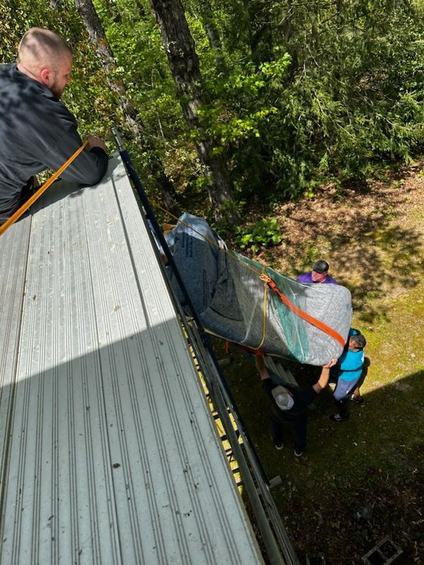 People loading a wrapped mattress onto a trailer from a rooftop. Bright sunlight, forest in the background.