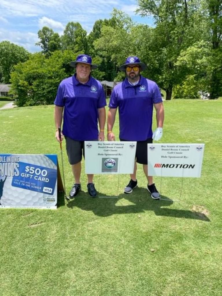 Two men in purple shirts pose on a golf course, holding signs for a golf tournament and a gift card.