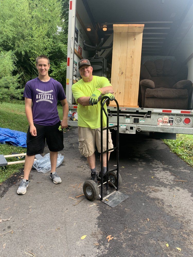 Two men loading furniture into a truck on a driveway. One man in a lime green shirt leans on a hand truck.