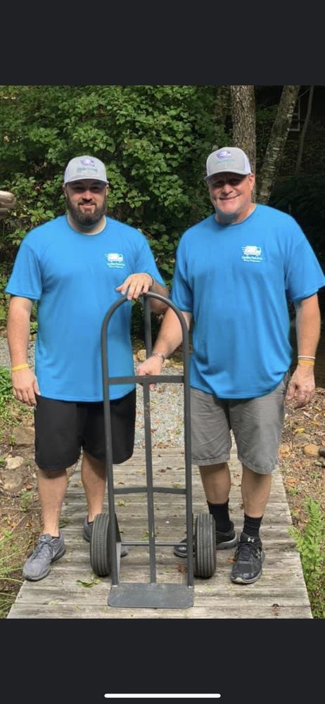 Two men in blue shirts and hats stand on a wooden path holding a hand truck.