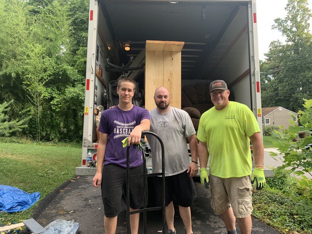 Three men loading a tall wooden cabinet into a truck. One in yellow, one in gray, and one in purple, smiling.
