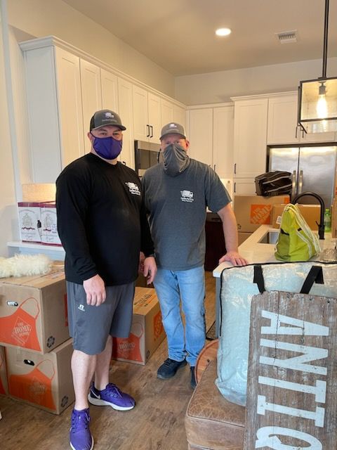 Two men in a kitchen wearing masks, surrounded by moving boxes. One in black shirt, other in gray.