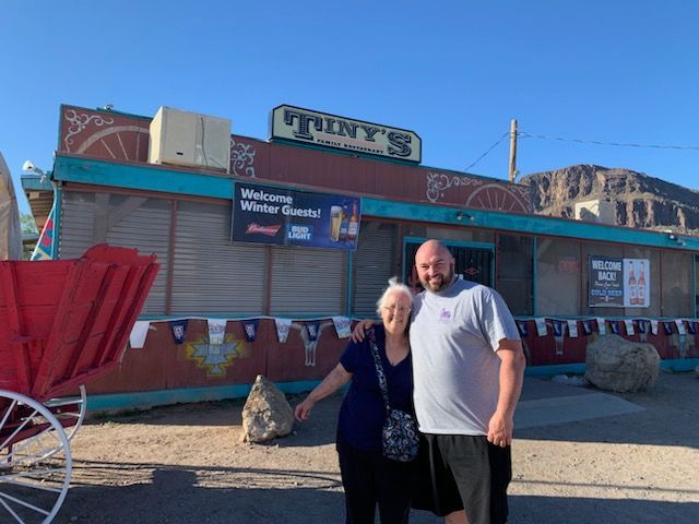 Two people pose in front of Tiny's Restaurant, a weathered Western-style building, blue sky backdrop.