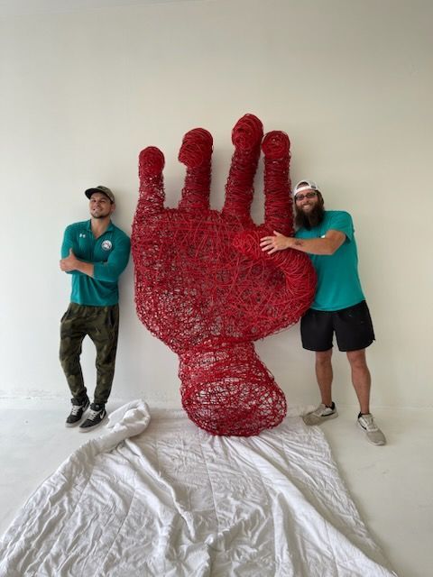 Two men pose beside a large, red hand sculpture in a white room.