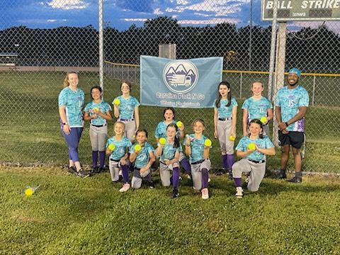Softball team posing on field in matching teal tie-dye shirts, gray pants, holding yellow balls.