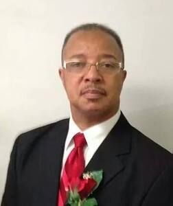 Man in a suit and tie, wearing glasses, and a red rose pinned on the lapel.