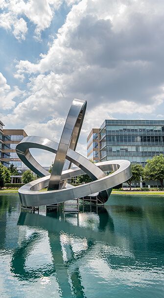 A large, abstract stainless steel sculpture on a pond, reflecting in the water; buildings in the background.