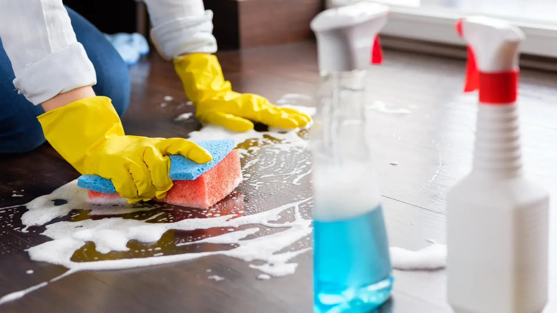 Person wearing yellow gloves cleaning a dark wood floor with a sponge and soapy water, with spray bottles in the background.
