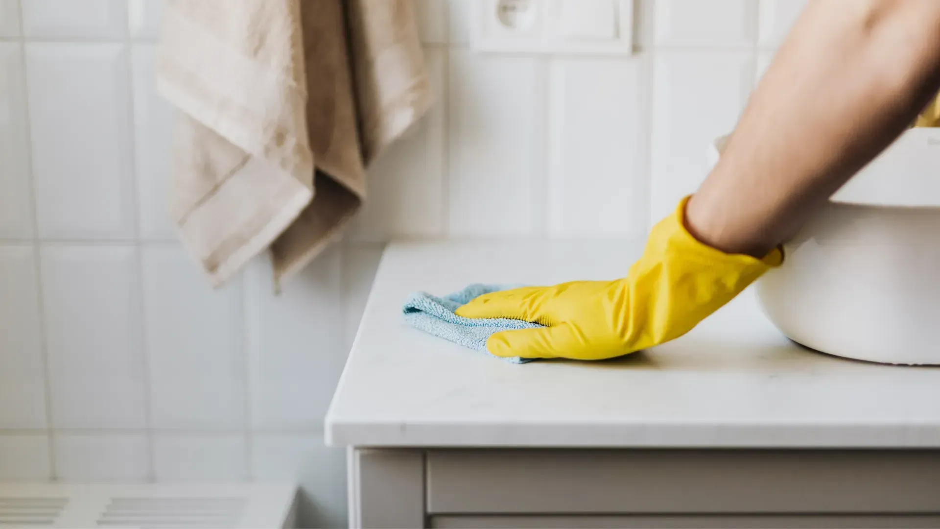 Person wearing a yellow glove wipes a white surface with a blue cloth in a bathroom. A towel hangs on the wall.