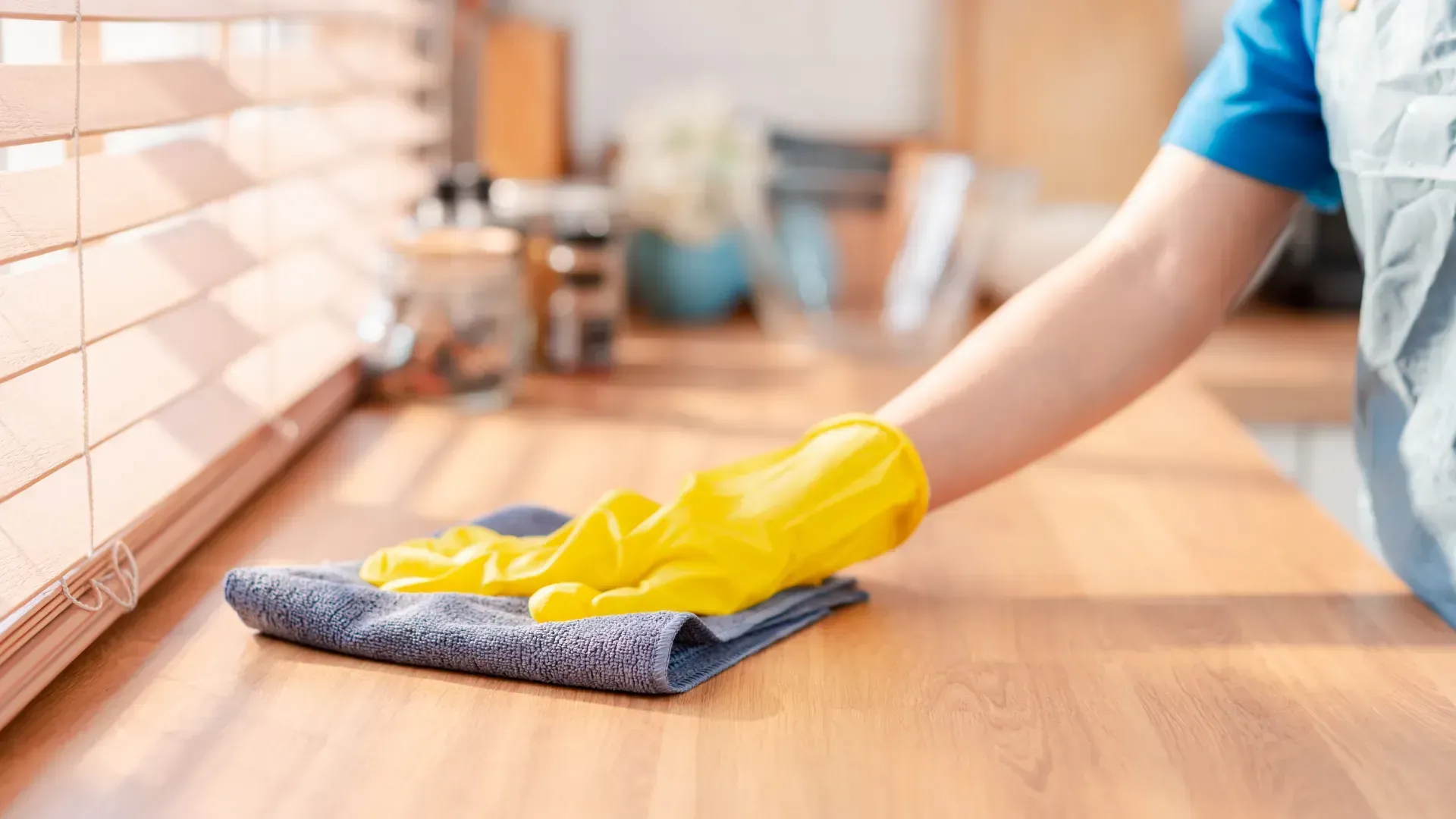 Person wearing yellow gloves wiping a wooden countertop with a gray cloth in a kitchen, near a window with blinds.