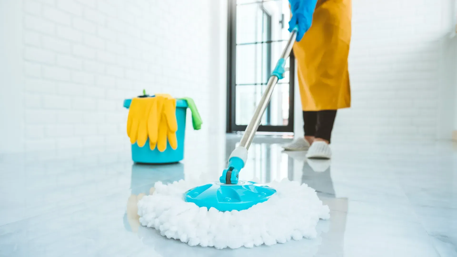 A person mops a white floor with a blue mop. A bucket with yellow gloves sits nearby.