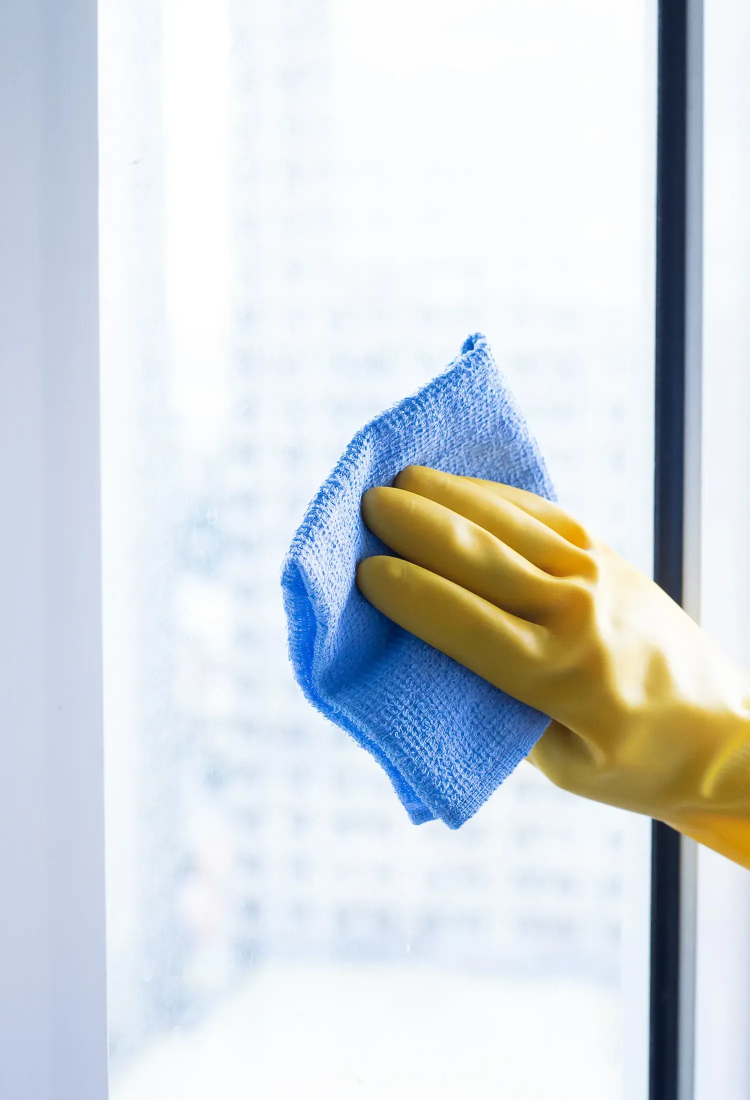 Yellow-gloved hand wiping a window with a blue microfiber cloth. The background is blurry, suggesting a cityscape.
