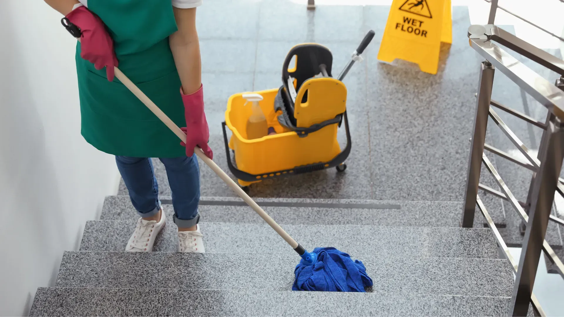 Person mopping stairs with a blue mop. A yellow mop bucket and wet floor sign are nearby.
