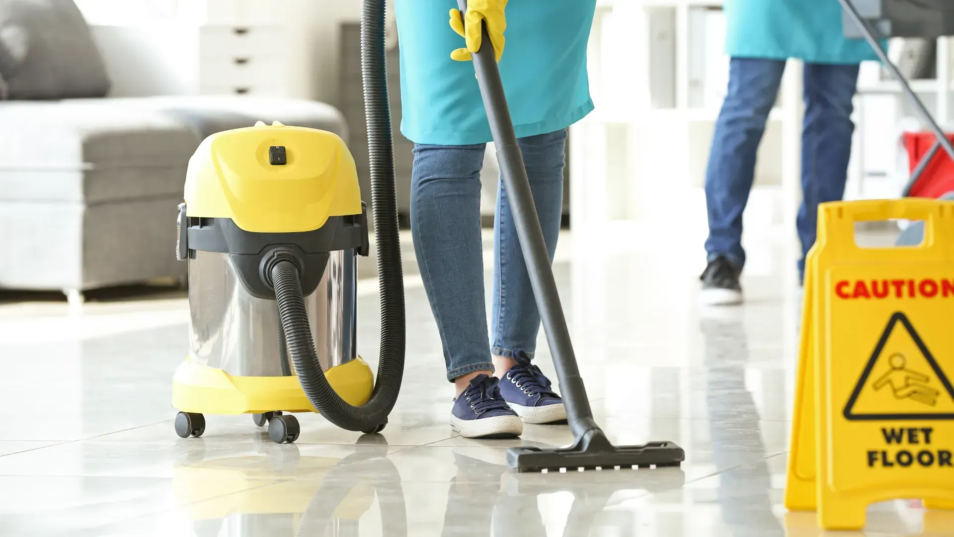 Two people cleaning a room; one vacuums with a yellow vacuum, the other stands nearby. A wet floor sign is present.