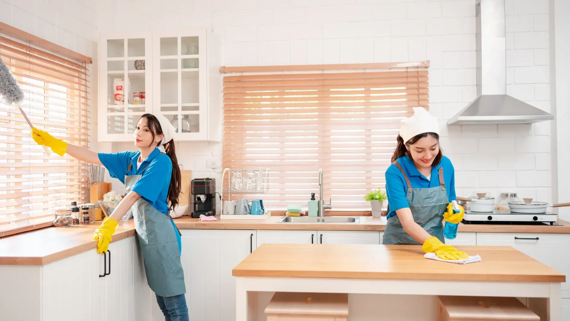 Two women cleaning a kitchen; one dusts a window, the other sprays and wipes a wooden table.