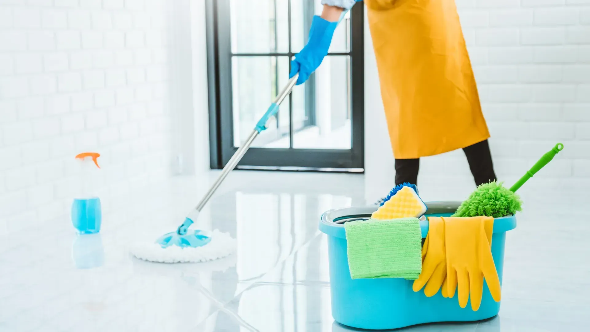 A person mopping a white floor with a blue mop and bucket, wearing gloves and an apron.