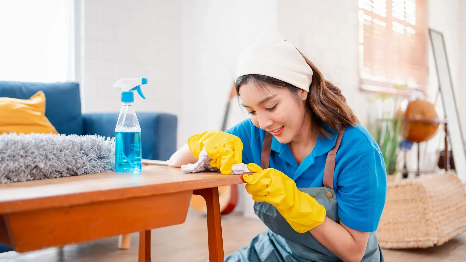 Woman wearing yellow gloves cleaning a wooden table in a brightly lit room with a blue couch.