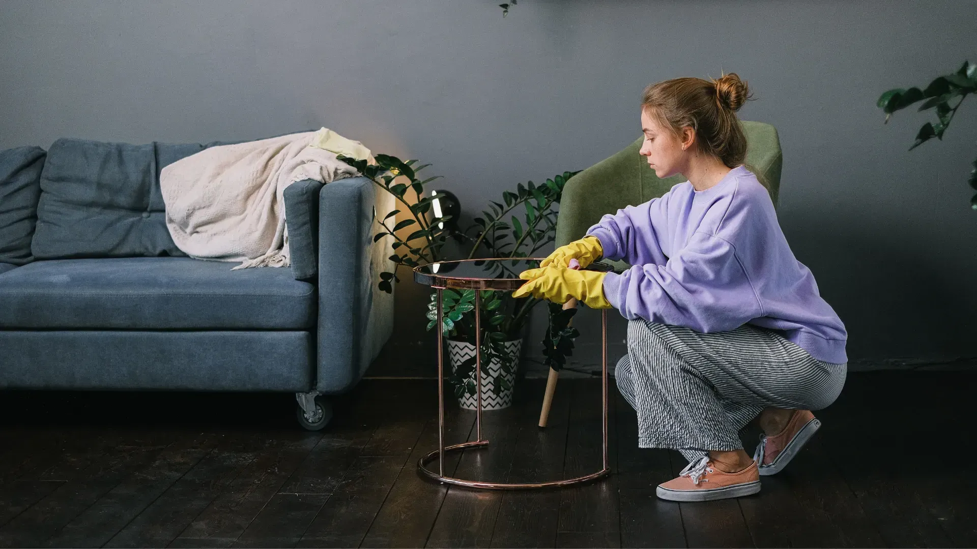 Woman wearing gloves squats, tending to a plant on a gold side table in a living room.