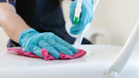 Person wearing blue gloves, cleaning a white chair with a pink cloth and spray bottle.