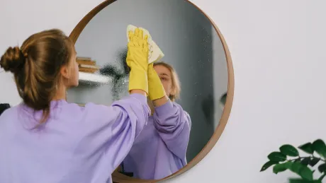 A woman wearing yellow gloves cleans a round mirror in a white room. She wears a purple sweatshirt, and her hair is in a bun.