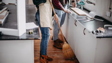 Person sweeping workshop floor while another works on a countertop.