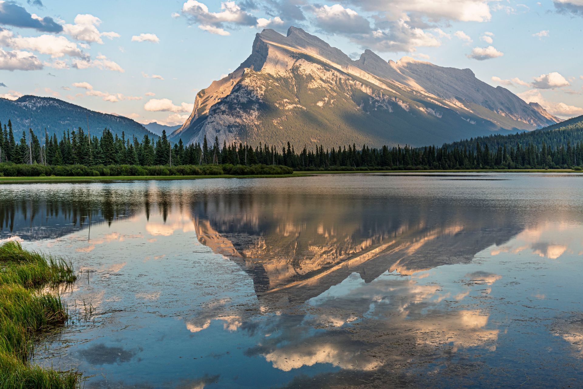 Mountain reflected in a calm lake; green trees line the shore, sky is partly cloudy.