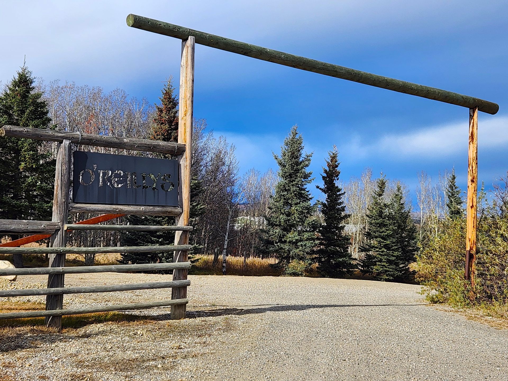 Rustic wooden entrance gate to a property, sign, trees, and gravel road under a blue sky.
