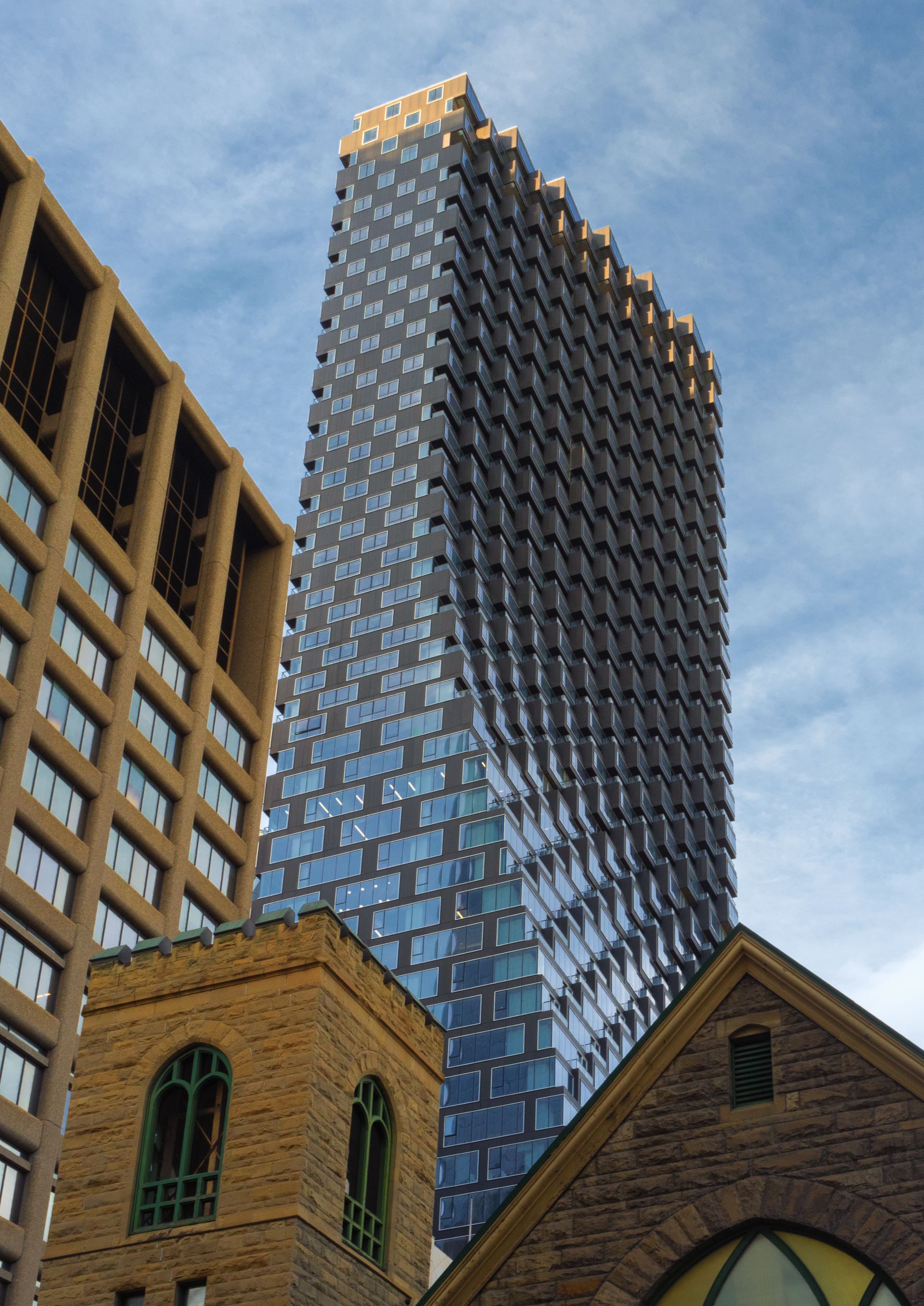 Skyscraper with patterned facade rises above a church and adjacent building against a blue sky.