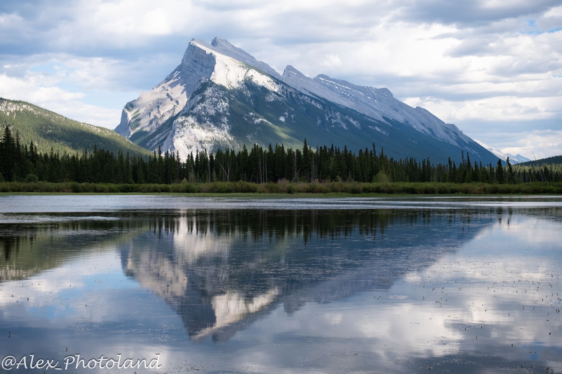 Snow-capped mountain reflecting in a calm lake, surrounded by green trees and cloudy sky.