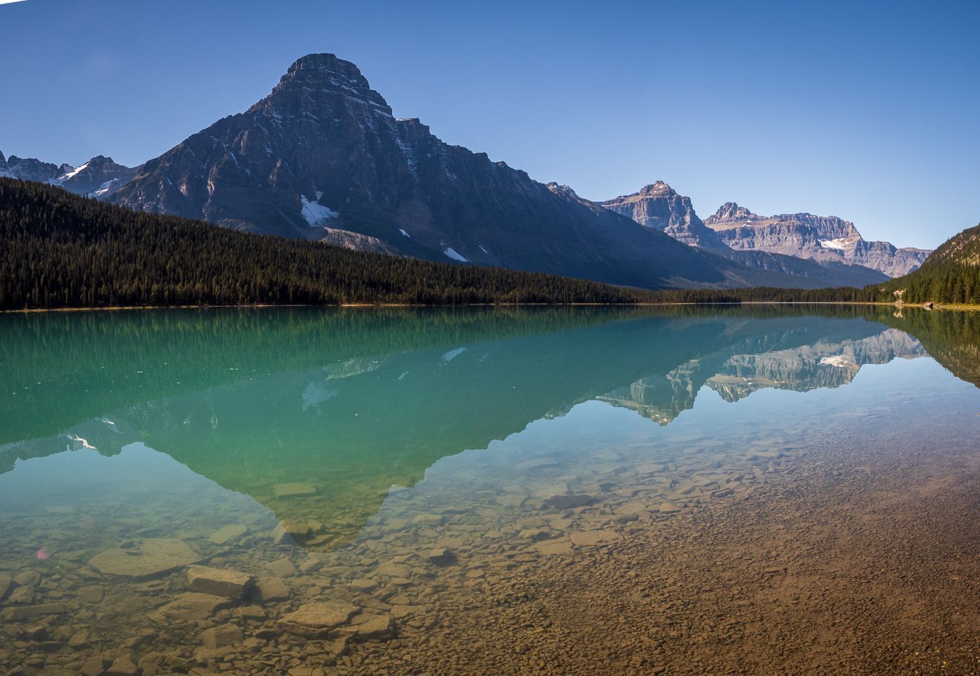 Emerald lake reflects a mountain and forest against a clear blue sky.