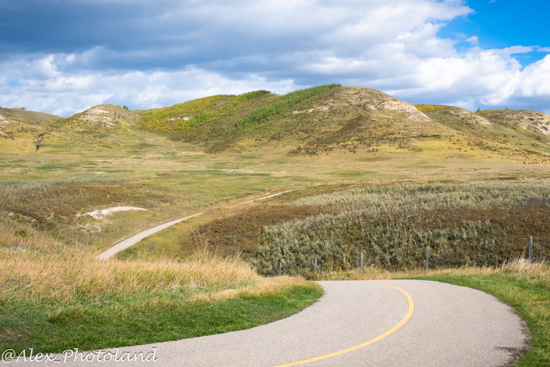Winding road through a grassy landscape with rolling hills under a cloudy blue sky.