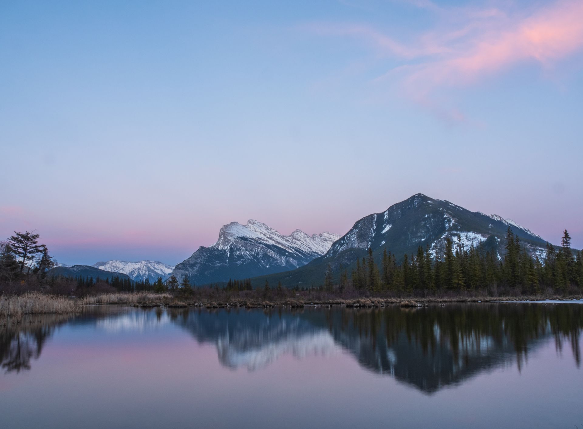 Mountains reflected in calm lake at dusk. Pink and purple sky, snow-capped peaks, evergreen trees.