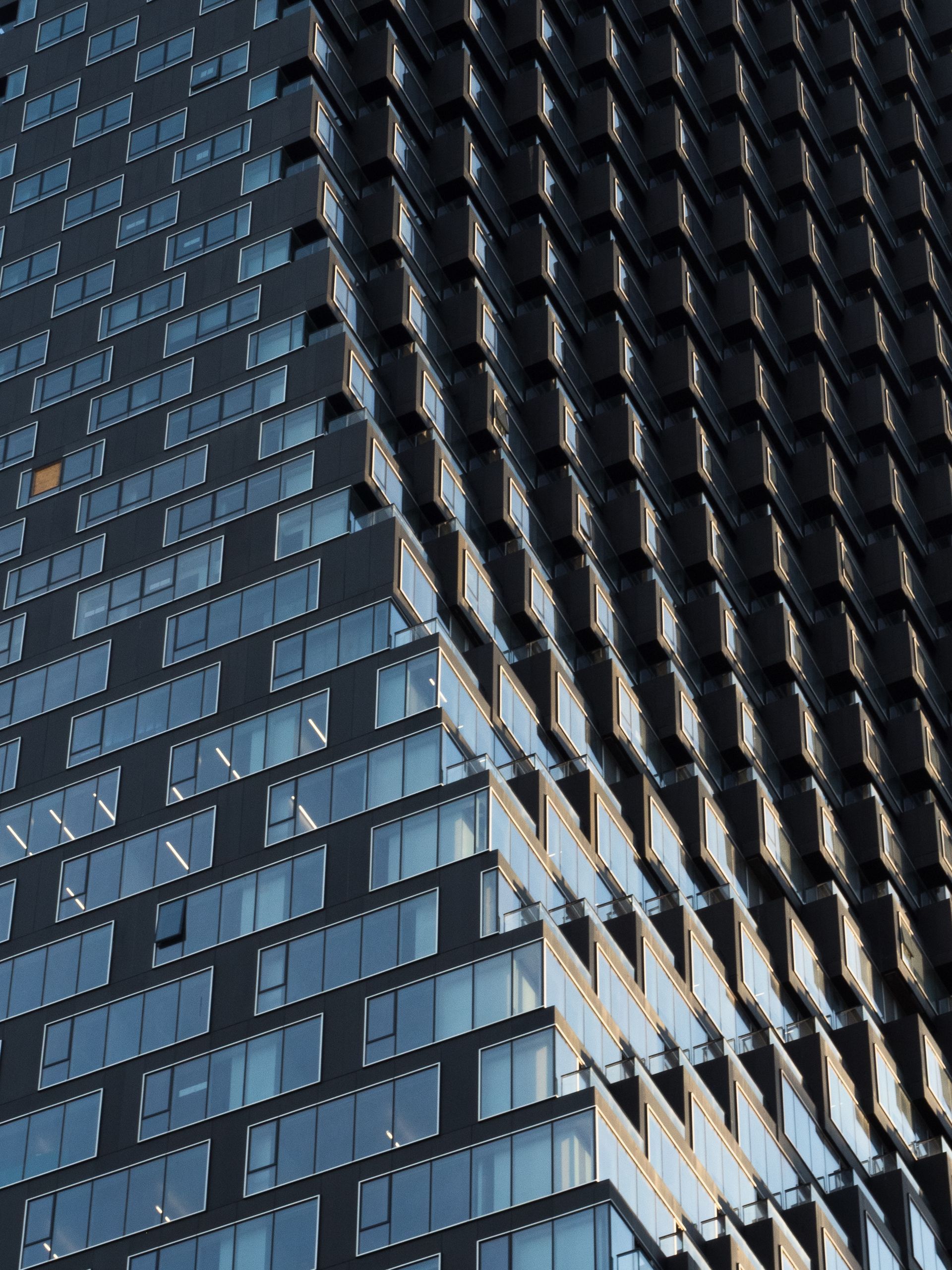 Modern skyscraper's corner, showcasing rows of windows and reflecting sunlight. Dark, geometric design.