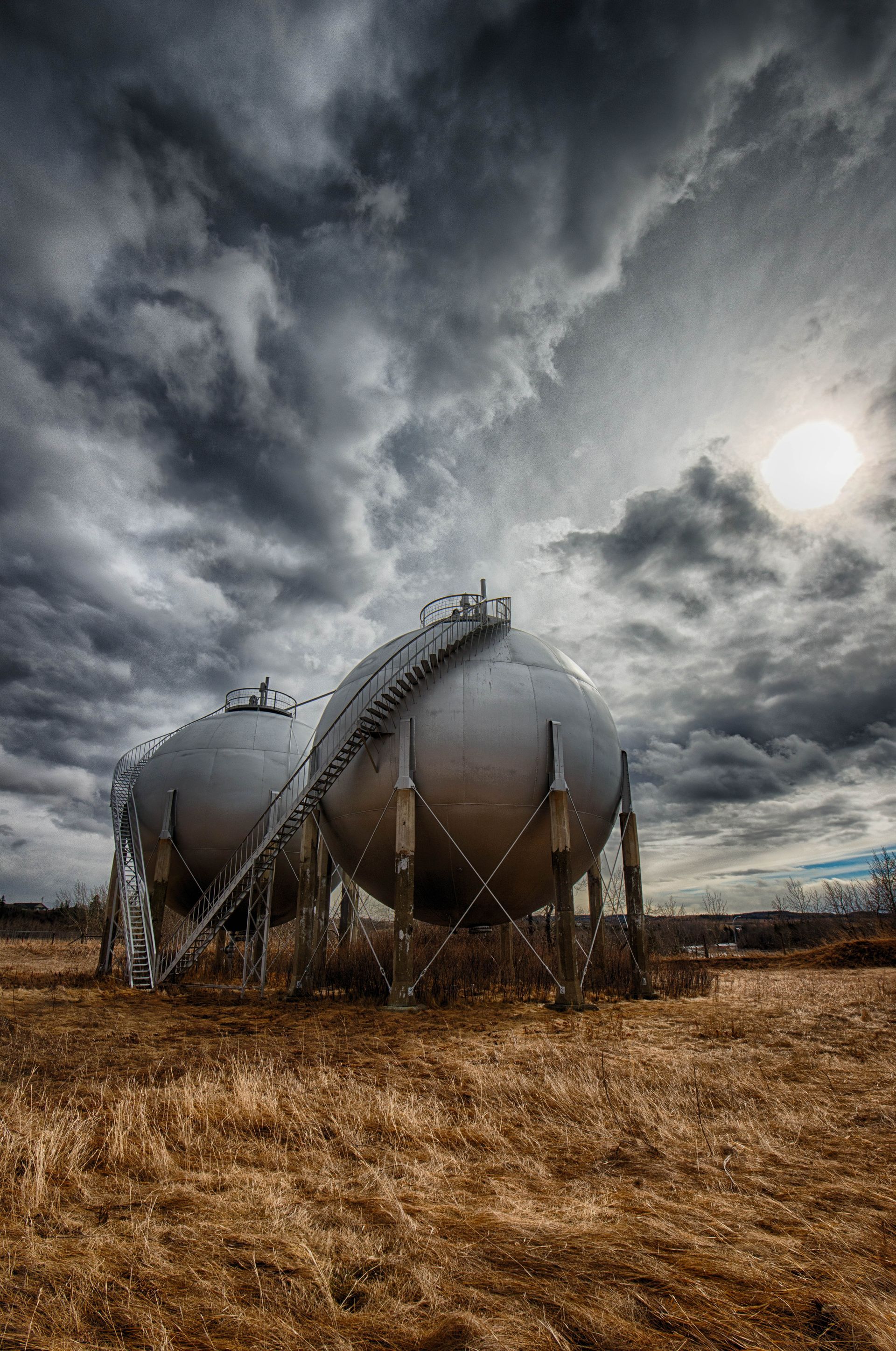 Two large spherical storage tanks under a stormy sky, in a field of brown grass.
